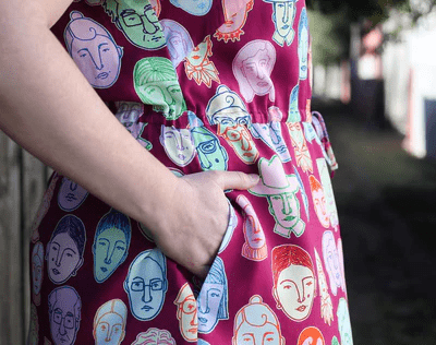 Woman in a bright red dress adorned with multi-colored abstract face patterns, standing confidently against a brick wall for a vibrant, artsy street-style vibe.