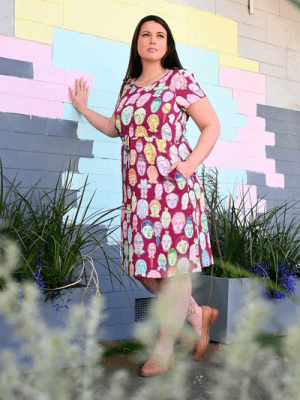 Woman in a bright red dress adorned with multi-colored abstract face patterns, standing confidently against a brick wall for a vibrant, artsy street-style vibe. Designed by Australian designer brand Streborclothing.