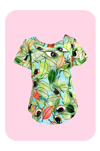 A short-sleeved top with a bold leaf and seed pattern in vibrant green, orange, and black on a light blue background, displayed against a pink backdrop to highlight its colorful appeal. Designed by Australian designer brand Streborclothing.