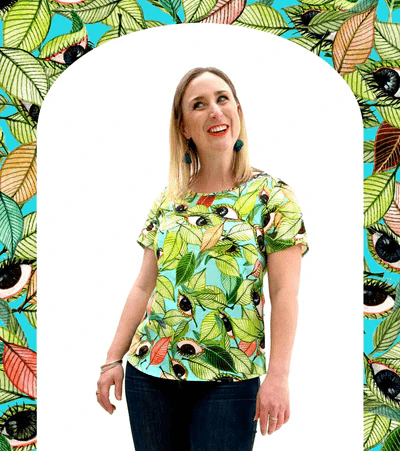 A short-sleeved top with a bold leaf and seed pattern in vibrant green, orange, and black on a light blue background, displayed against a pink backdrop to highlight its colorful appeal. Designed by Australian designer brand Streborclothing.