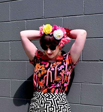 Woman wearing a vibrant black top with a pink and orange desert-themed print, posed against a similarly themed background, emphasizing the bold, artistic nature of Strebor's style. Designed by Australian designer brand Streborclothing.