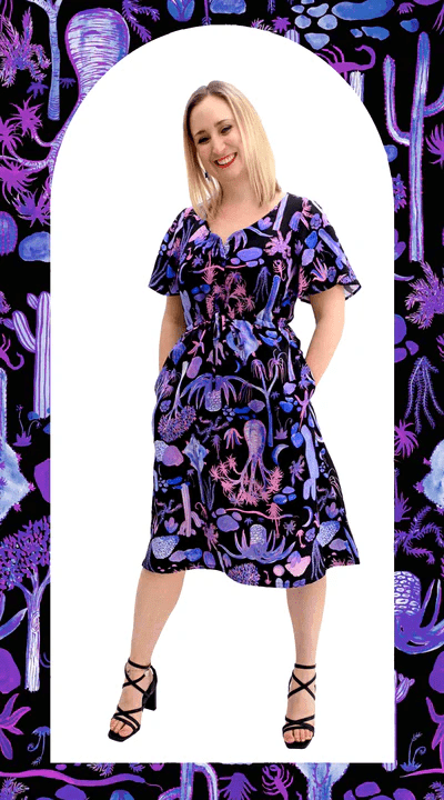 Woman wearing a dark dress with a whimsical cactus and desert-themed print in pinks, purples, and blues, standing against a graphic backdrop with matching patterns. Designed by Australian designer brand Streborclothing.