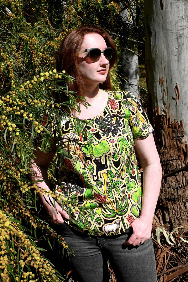This top showcases a complex pattern with elements of flora, including leaves, flowers, and abstract natural shapes in shades of green, red, and yellow. The design appears dense and rich, resembling an intricate forest scene. Designed by Australian designer brand Streborclothing.