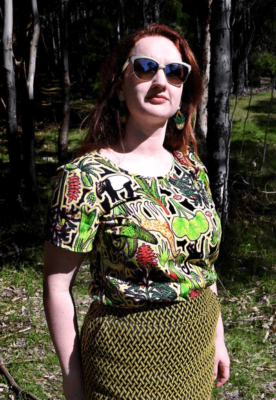 This top showcases a complex pattern with elements of flora, including leaves, flowers, and abstract natural shapes in shades of green, red, and yellow. The design appears dense and rich, resembling an intricate forest scene. Designed by Australian designer brand Streborclothing.