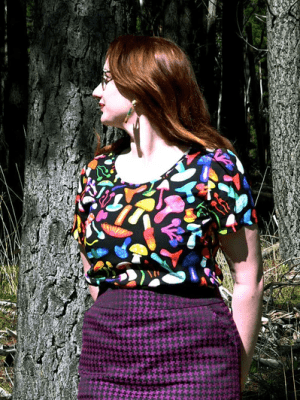 The dress features a vibrant mushroom pattern with various brightly colored mushrooms in red, yellow, blue, pink, and green on a black background. This playful design adds a whimsical and organic feel to the outfit. Designed by Australian designer brand Streborclothing.